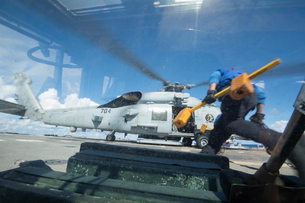 A U.S. Navy helicopter, numbered 704, sits on what appears to be an aircraft carrier flight deck with its rotors spinning. The image is shot through a glass windshield or window, creating reflections and distortion. A crew member in a blue shirt is visible in motion, carrying yellow chocks or safety equipment.
