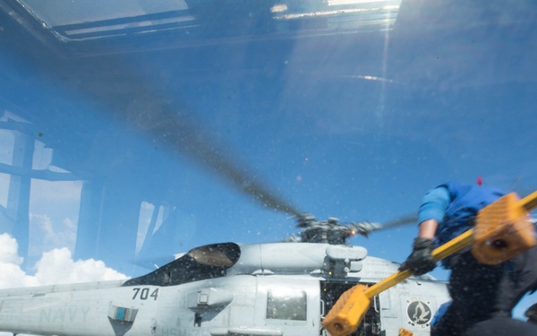 A U.S. Navy helicopter, numbered 704, sits on what appears to be an aircraft carrier flight deck with its rotors spinning. The image is shot through a glass windshield or window, creating reflections and distortion. A crew member in a blue shirt is visible in motion, carrying yellow chocks or safety equipment.