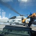 A U.S. Navy helicopter, numbered 704, sits on what appears to be an aircraft carrier flight deck with its rotors spinning. The image is shot through a glass windshield or window, creating reflections and distortion. A crew member in a blue shirt is visible in motion, carrying yellow chocks or safety equipment.