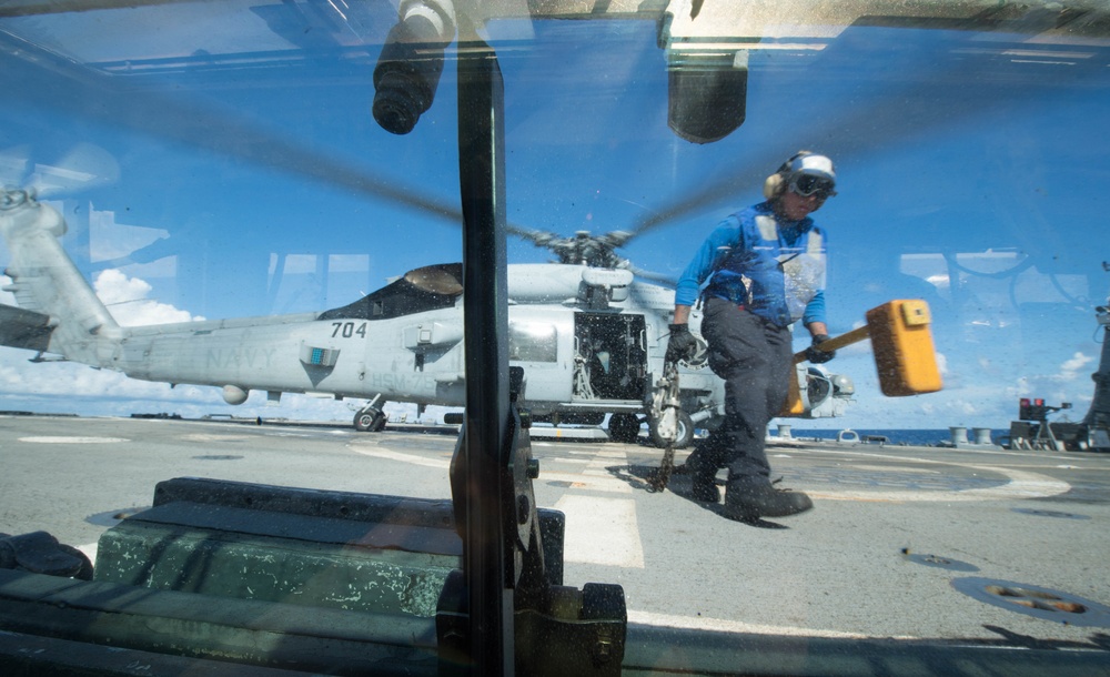 The image is taken through a vehicle or equipment windshield on what appears to be a naval vessel flight deck. A deck crew member wearing a blue shirt, protective gear, and helmet is carrying a large yellow mallet or chock tool near a military helicopter marked '704'. The helicopter appears to be an MH-60 Seahawk variant with rotors spinning, suggesting active flight operations.
