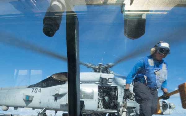 The image is taken through a vehicle or equipment windshield on what appears to be a naval vessel flight deck. A deck crew member wearing a blue shirt, protective gear, and helmet is carrying a large yellow mallet or chock tool near a military helicopter marked '704'. The helicopter appears to be an MH-60 Seahawk variant with rotors spinning, suggesting active flight operations.