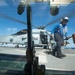 The image is taken through a vehicle or equipment windshield on what appears to be a naval vessel flight deck. A deck crew member wearing a blue shirt, protective gear, and helmet is carrying a large yellow mallet or chock tool near a military helicopter marked '704'. The helicopter appears to be an MH-60 Seahawk variant with rotors spinning, suggesting active flight operations.