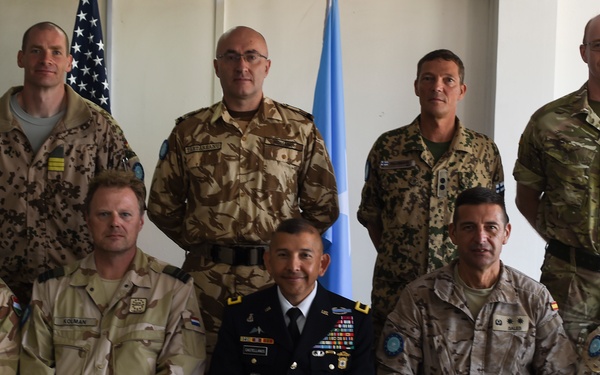 A formal group photograph of nine military personnel in a posed arrangement with four standing in the back row and five seated in the front row. The individuals are wearing various national military uniforms, suggesting a multinational military gathering or meeting. An American flag and a United Nations flag are visible in the background.