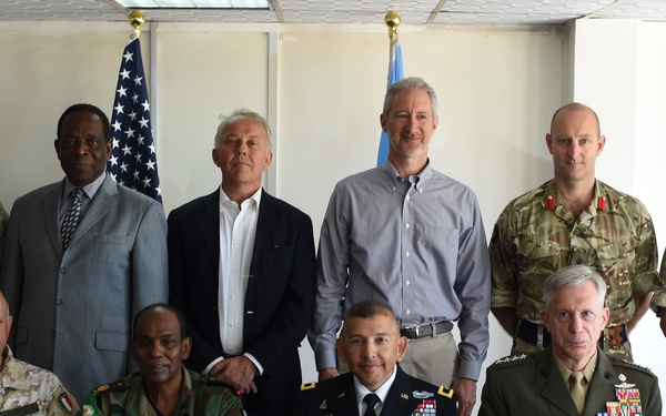 A formal group photograph of approximately eleven individuals, a mix of military personnel in various uniforms and civilians, posed in two rows in an indoor setting. Two flags are visible in the background — an American flag and what appears to be a United Nations flag. The gathering appears to be a multinational military or diplomatic meeting.