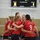A women's volleyball team wearing red 'Deutschland' (Germany) jerseys huddles together on an indoor court, likely during a match. The players are grouped in a tight circle with arms around each other, suggesting a team moment such as celebrating a point or strategizing. A scoreboard and volleyball net are visible in the background.