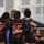 A group of female volleyball players wearing black and red 'Canada' jerseys are huddled together in a team huddle, with their arms around each other's shoulders. Visible jersey numbers include 15, 9, and a partially visible number on the leftmost player. The setting appears to be an indoor volleyball court with a net visible in the background.