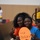 Two young women are posing together and smiling at the camera in what appears to be an indoor gymnasium or sports facility. One is wearing an orange athletic jersey and the other a blue t-shirt, and they are holding an orange oval-shaped sign that reads 'Ga Holland' with a windmill illustration. They appear to be spectators or supporters at a sporting event.