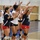 A women's volleyball team wearing USA uniforms in white and navy blue with red shorts is celebrating on an indoor court. The players are clapping, raising their arms, and smiling, suggesting a positive moment such as a point scored or a game won. A volleyball net is visible in the background of what appears to be an indoor gymnasium.
