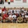 A large group photo of a women's volleyball team wearing USA uniforms, along with coaches and support staff, posing together on an indoor volleyball court. The scoreboard in the background shows Period 2 with a 3:00 time display. Several national flags, including Germany and the USA, are visible on the walls in the background.