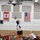 A volleyball match is being played in an indoor gymnasium identified as a Navy Fitness facility. A player wearing number 6 in a white uniform is jumping to spike or set the ball near the net, while teammates and opponents in red uniforms are positioned around the court. Two national flags — Canadian and American — are displayed on the back wall above the court.