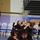 A group of female volleyball players wearing black and red 'Canada' uniforms are celebrating together in a huddle on an indoor court. At least two jersey numbers are visible: #9 and #15. The players are embracing and raising their hands in celebration, suggesting a successful play or point scored.