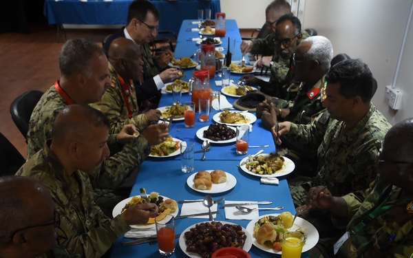 A group of military personnel from multiple nations are seated at a long rectangular table covered with a blue tablecloth, sharing a formal meal together. The attendees are wearing various military uniforms and camouflage patterns, suggesting a multinational military gathering or conference luncheon. The table is set with multiple food dishes, beverages including orange juice, and standard dining utensils.