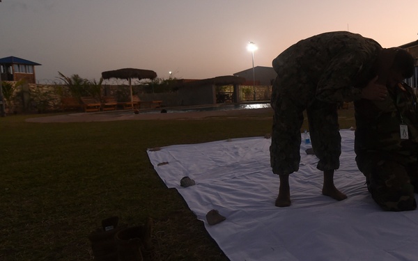 Two individuals in military camouflage uniforms are standing barefoot on a white sheet or tarp spread on a grass lawn at dusk. Multiple pairs of boots have been removed and placed on the grass beside the tarp. The setting appears to be a military compound or base facility with a pool, perimeter wall, and lit buildings visible in the background.