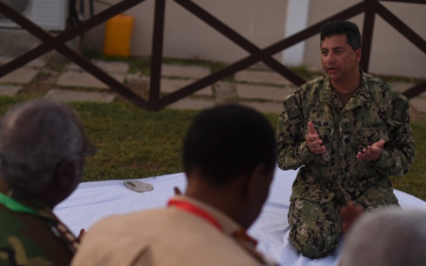 A US Navy servicemember in digital camouflage uniform is kneeling on a white cloth or tarp, appearing to conduct a briefing or demonstration for a group of individuals in various military uniforms. The setting appears to be an outdoor courtyard or compound area. The audience members are seated or kneeling facing the presenter, suggesting a training or instructional session.