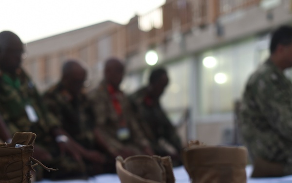 The image shows a group of military personnel in various camouflage uniforms kneeling or prostrating on a white mat or cloth outdoors, suggesting they are engaged in prayer. In the foreground, several pairs of tan/brown military boots have been removed and placed on the grass, consistent with Islamic prayer practice of removing footwear. The scene appears to take place at dusk or dawn near a building with lights visible in the background.