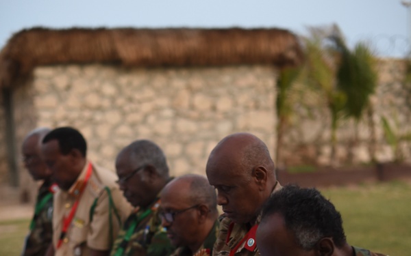 A group of military officers in various camouflage uniforms are kneeling in prayer on a white cloth outdoors. The rightmost individual wears a patch identifiable as the Somali flag on his left shoulder. Military caps have been removed and placed on the ground, consistent with Islamic prayer practice.