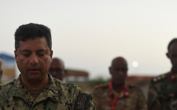 A U.S. Navy servicemember named Saifulislam, wearing NWU Type III camouflage with U.S. flag patches, stands with arms crossed and eyes closed in what appears to be a prayer or moment of reflection. Several other military personnel in various uniforms, likely from African nations, stand in the background in similar postures. The setting appears to be an outdoor military installation at dusk or dawn.