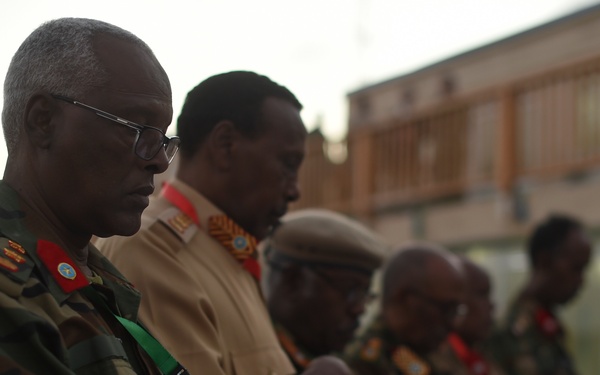 A group of military officers in uniform are standing in a line, appearing to be at attention or in a moment of reflection, possibly during a ceremony or formal gathering. The foreground figure wears camouflage fatigues with rank insignia, glasses, a gold watch, and carries a small notebook with a green lanyard. The setting appears to be an outdoor area near a building, photographed in low ambient light conditions.
