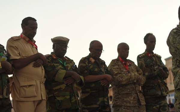 A group of military personnel in various uniforms are standing in a line on a white mat or cloth, with arms crossed or folded, appearing to be engaged in a prayer or moment of reflection. The group includes individuals in different camouflage patterns and one person in a tan/khaki dress uniform, suggesting a multinational military gathering. One individual on the far right wears what appears to be a US Navy working uniform (NWU).
