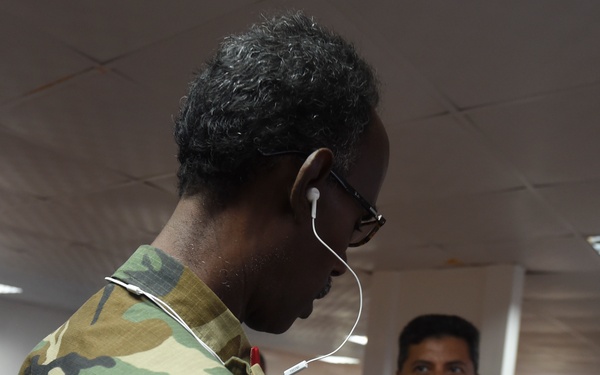 Two military personnel in different camouflage uniforms are interacting indoors. The foreground figure wears woodland camouflage and has a white earbud in their ear connected to a mobile device being held at chest level. A second individual in a different camouflage pattern (appears to be US Navy NWU or similar digital pattern) stands in the background, appearing to speak or gesture.
