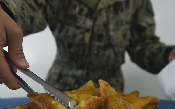 A person wearing a military camouflage uniform (consistent with U.S. Navy Working Uniform) is using metal tongs to serve samosas from a large silver/stainless steel platter. The samosas appear golden-brown and freshly fried, piled on the plate atop a blue tablecloth. The person's other hand holds what appears to be a white napkin or paper.