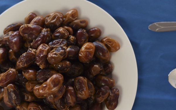 A large white plate is piled high with dates (the fruit), placed on a blue tablecloth. To the right of the plate, a white napkin holds a knife, fork, and spoon arranged neatly. The image is taken from a top-down perspective with good lighting.