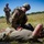 A U.S. Army soldier in full combat uniform and gear is performing tactical combat casualty care (TCCC) on another soldier lying on the ground in an open field. A third soldier stands in the background appearing to observe or evaluate the exercise, holding what appears to be a notepad or clipboard. This appears to be a field medical training exercise based on the controlled setting and evaluator presence.