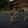 Three military personnel in camouflage uniforms are engaged in hand-to-hand combat training on a rubber-surfaced outdoor area. Two soldiers are actively sparring in the foreground while a third observes or waits in the background. The setting appears to be a military installation with brick barracks buildings visible in the background.