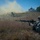 Two soldiers in camouflage uniforms are in prone firing positions in a grassy field during what appears to be a military training exercise. A large dust cloud is visible in the background, likely from a simulated explosion or smoke device. A building with a blue dome is visible in the background, suggesting a mock urban environment used for training.