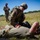 A U.S. Army soldier in full combat uniform and gear is performing tactical combat casualty care (TCCC) on another soldier lying on the ground in an open field. A third soldier stands in the background appearing to observe and take notes, suggesting this is a training exercise or evaluation. The scene takes place outdoors in a grassy field under clear blue skies.