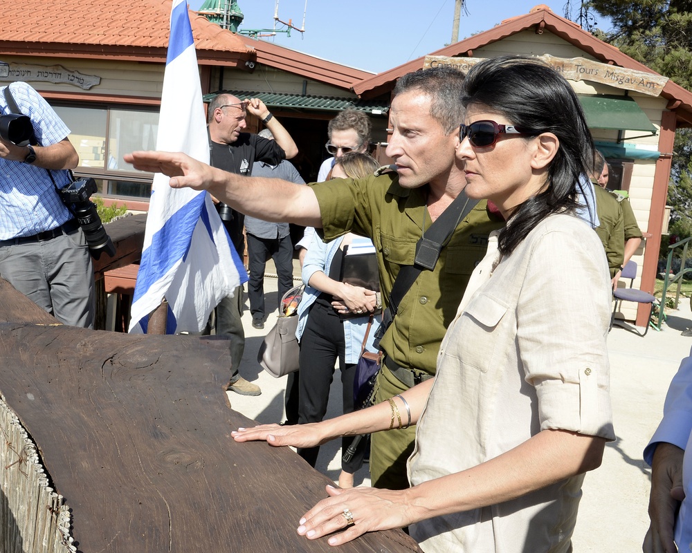 A group of people are gathered outdoors near a building with a sign reading 'Tours Misgav Am' and Hebrew text. A person in Israeli military uniform is pointing toward something in the distance while standing next to a woman in a beige outfit wearing sunglasses. An Israeli flag is visible in the background along with photographers and other attendees.