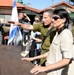 A group of people are gathered outdoors near a building with a sign reading 'Tours Misgav Am' and Hebrew text. A person in Israeli military uniform is pointing toward something in the distance while standing next to a woman in a beige outfit wearing sunglasses. An Israeli flag is visible in the background along with photographers and other attendees.