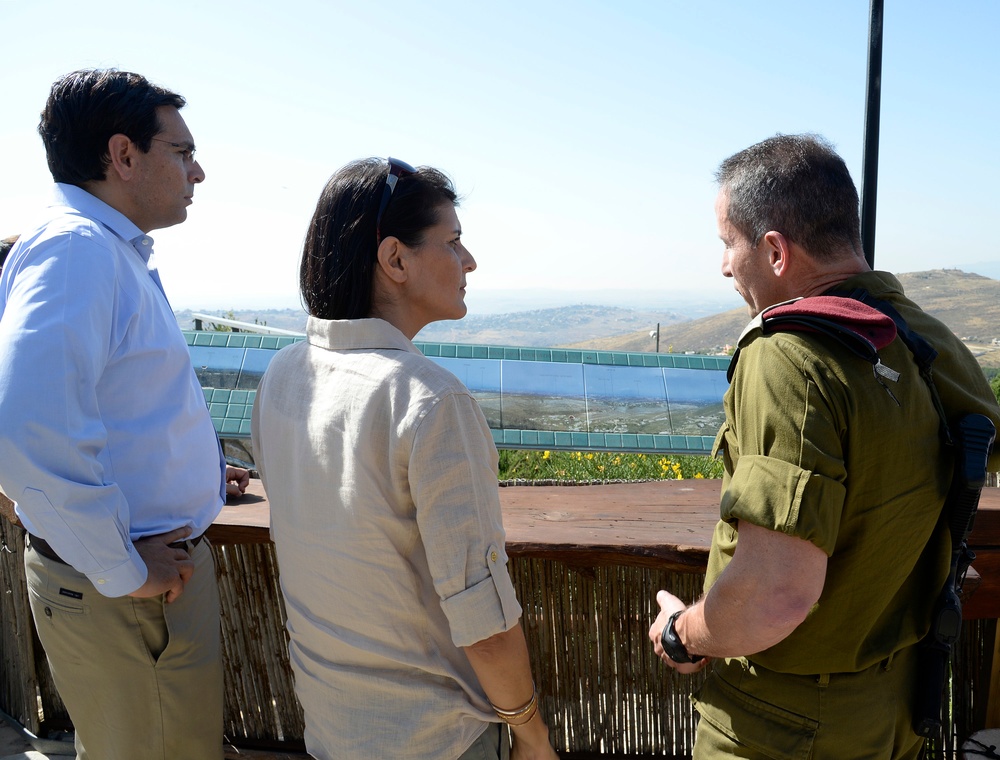Three individuals are standing on an elevated outdoor terrace or observation point, engaged in conversation. Two civilians (one man, one woman) are speaking with a person in military uniform, with a scenic landscape featuring a body of water and hills visible in the background. The setting appears to be a formal or official visit to a strategic overlook.