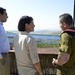 Three individuals are standing on an elevated outdoor terrace or observation point, engaged in conversation. Two civilians (one man, one woman) are speaking with a person in military uniform, with a scenic landscape featuring a body of water and hills visible in the background. The setting appears to be a formal or official visit to a strategic overlook.