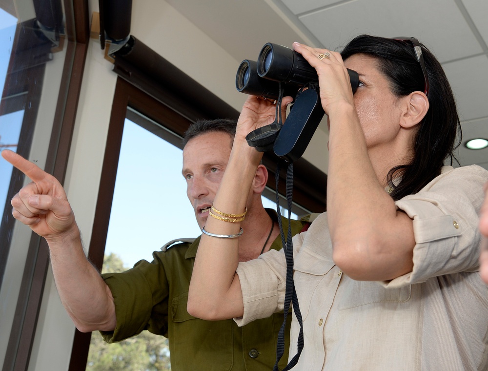 Two individuals are inside a building near a window, with one person looking through black binoculars while the other points in a direction, apparently guiding the observation. The man is wearing an olive/military-style green shirt, while the woman is dressed in a beige/khaki shirt with sunglasses resting on her head. The scene suggests a briefing or guided observation activity.