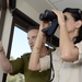 Two individuals are inside a building near a window, with one person looking through black binoculars while the other points in a direction, apparently guiding the observation. The man is wearing an olive/military-style green shirt, while the woman is dressed in a beige/khaki shirt with sunglasses resting on her head. The scene suggests a briefing or guided observation activity.