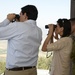 Three individuals are standing at an elevated observation point overlooking a wide valley or agricultural landscape. Two civilians, a man and a woman, are using binoculars to survey the terrain below, while a uniformed military officer stands beside them. The setting appears to be a military or official observation post based on the structure and the presence of the uniformed personnel.