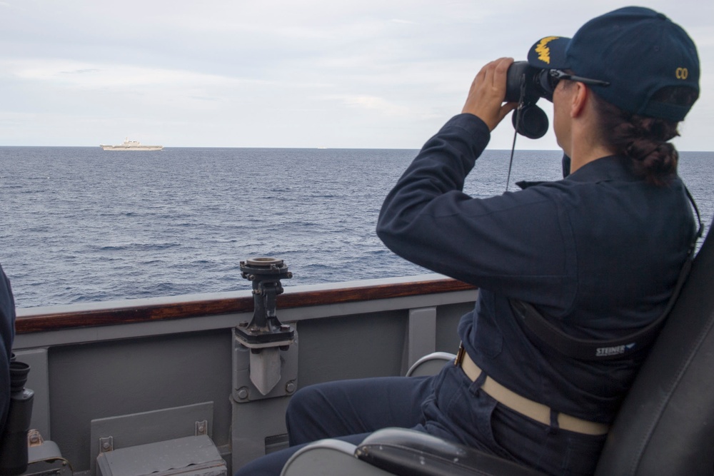 A naval officer wearing a dark blue uniform and a cap marked 'CO' (Commanding Officer) is seated on a ship's bridge wing, observing a distant vessel through Steiner binoculars. The distant ship on the horizon appears to be a large naval vessel, possibly an aircraft carrier or amphibious assault ship. The scene takes place on calm open ocean under overcast skies.