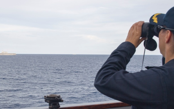A naval officer wearing a dark blue uniform and a cap marked 'CO' (Commanding Officer) is seated on a ship's bridge wing, observing a distant vessel through Steiner binoculars. The distant ship on the horizon appears to be a large naval vessel, possibly an aircraft carrier or amphibious assault ship. The scene takes place on calm open ocean under overcast skies.