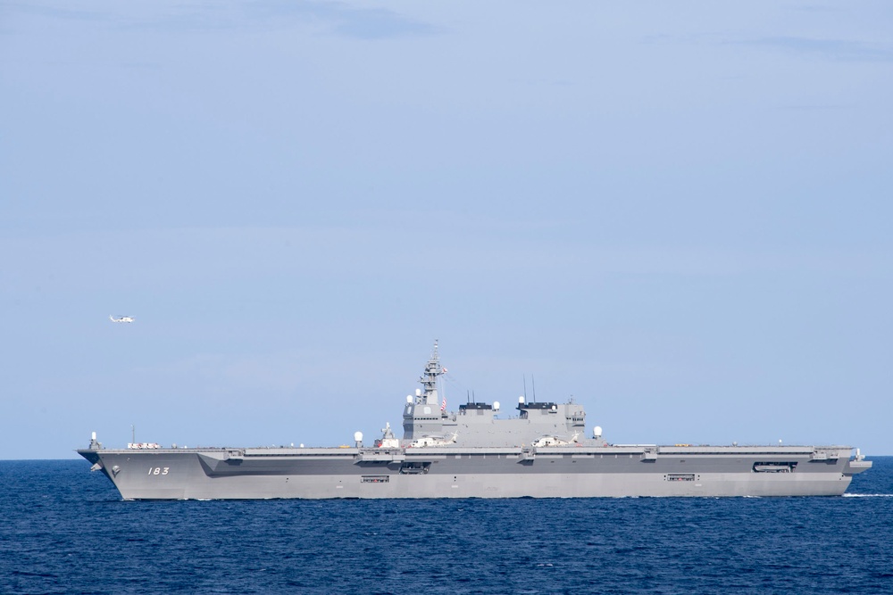 A large gray flat-deck warship bearing hull number 183 is underway on open blue ocean waters. The vessel appears to be a helicopter destroyer or light carrier-type ship of the Japan Maritime Self-Defense Force (JMSDF), identifiable as JS Izumo (DDH-183). A small aircraft or helicopter is visible in the background sky to the left of the ship.