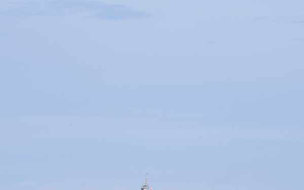 A large gray flat-deck warship bearing hull number 183 is underway on open blue ocean waters. The vessel appears to be a helicopter destroyer or light carrier-type ship of the Japan Maritime Self-Defense Force (JMSDF), identifiable as JS Izumo (DDH-183). A small aircraft or helicopter is visible in the background sky to the left of the ship.