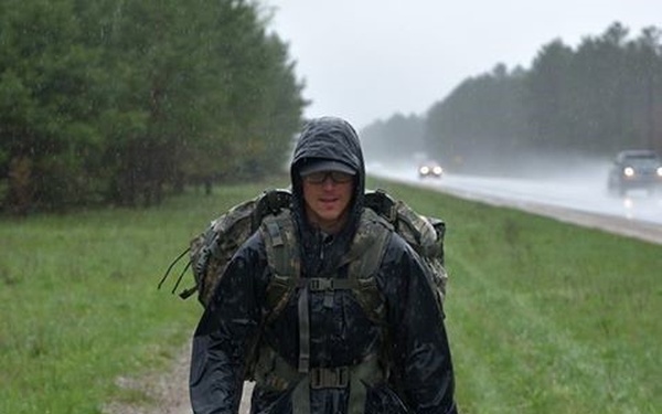 A person is walking along a wet gravel shoulder of a highway during heavy rain. They are carrying a large military-style camouflage backpack and wearing a dark rain jacket with the hood up, shorts, compression socks, and running shoes. The background shows a rain-soaked road with vehicle headlights visible in the distance and tree lines on both sides.