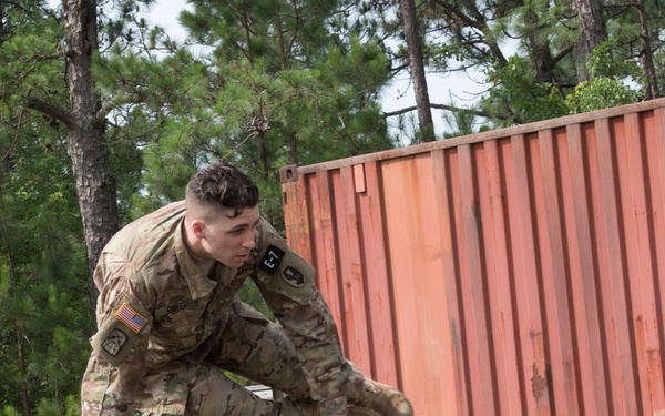 A U.S. Army soldier in MultiCam/OCP camouflage uniform is navigating an obstacle course, appearing to vault or jump over a wooden beam structure. A second soldier in ACU (Army Combat Uniform) pattern observes from the right side. The setting appears to be a military training facility with a shipping container and chain-link fence visible in the background.