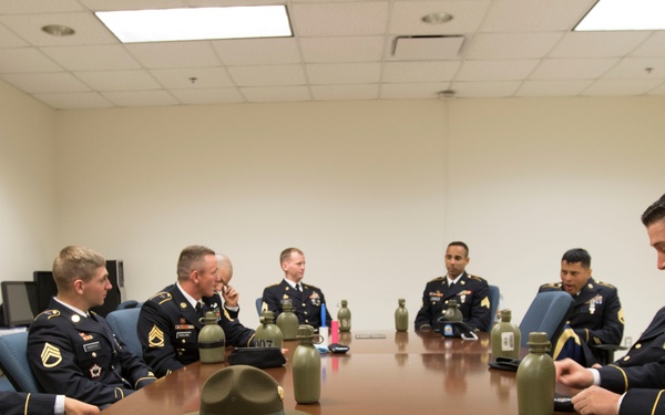 A group of U.S. Army soldiers in dress blue uniforms are seated around a large conference table in a formal meeting room. Multiple ranks are visible including sergeants and staff sergeants. A campaign/drill sergeant hat and several military canteens are placed on the table as centerpieces.