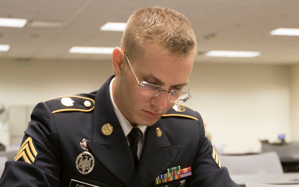 A young U.S. Army soldier in dress blues uniform is seated at a desk writing on a document. His name tape reads 'DENNIS' and he wears sergeant chevrons along with several service ribbons and a badge. The setting appears to be a military classroom or administrative office.