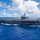 170612-N-BL637-168 A large US Navy aircraft carrier is underway in open blue ocean under partly cloudy skies. The flight deck is populated with numerous aircraft arranged along its edges. The vessel is moving at speed, producing visible wake on both sides of the hull.