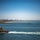 170613-N-TV230-163 A red tugboat numbered '11' navigates through a harbor channel, leaving a white wake behind it. In the background, a city skyline is visible across the bay, along with a long bridge, a sandy beach, and a rock jetty. The scene appears to be a busy port entrance under clear blue skies.