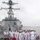 Sterett-Dewey Surface Action Group Deployment A group of naval personnel in white dress uniforms are gathered on the deck of a gray warship, appearing to be engaged in a formal visit or tour. The ship features a prominent superstructure with radar domes, a forward gun mount, and various naval equipment. The setting appears to be a port or harbor with calm water and an overcast sky visible in the background.