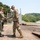 170615-N-NY430-0117 Two military personnel in different uniforms are shaking hands on a concrete pier or dock area near a waterway. One individual wears U.S. Navy Working Uniform (NWU) while the other wears Army Combat Uniform (ACU/OCP multicam pattern). A third person in civilian attire is visible in the background near a chain-link security fence.