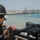 170614-N-FQ994-191 A U.S. Navy sailor in tactical gear mans a mounted machine gun aboard a vessel in a harbor or port area. The sailor is wearing a ballistic helmet, sunglasses, and a body armor vest with 'US NAVY' markings. In the background, multiple naval warships are visible docked at what appears to be a military port facility.
